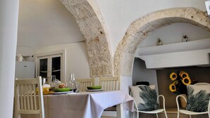 Un'area pranzo con un tavolo apparecchiato per due, sedie bianche e un arco in pietra.
