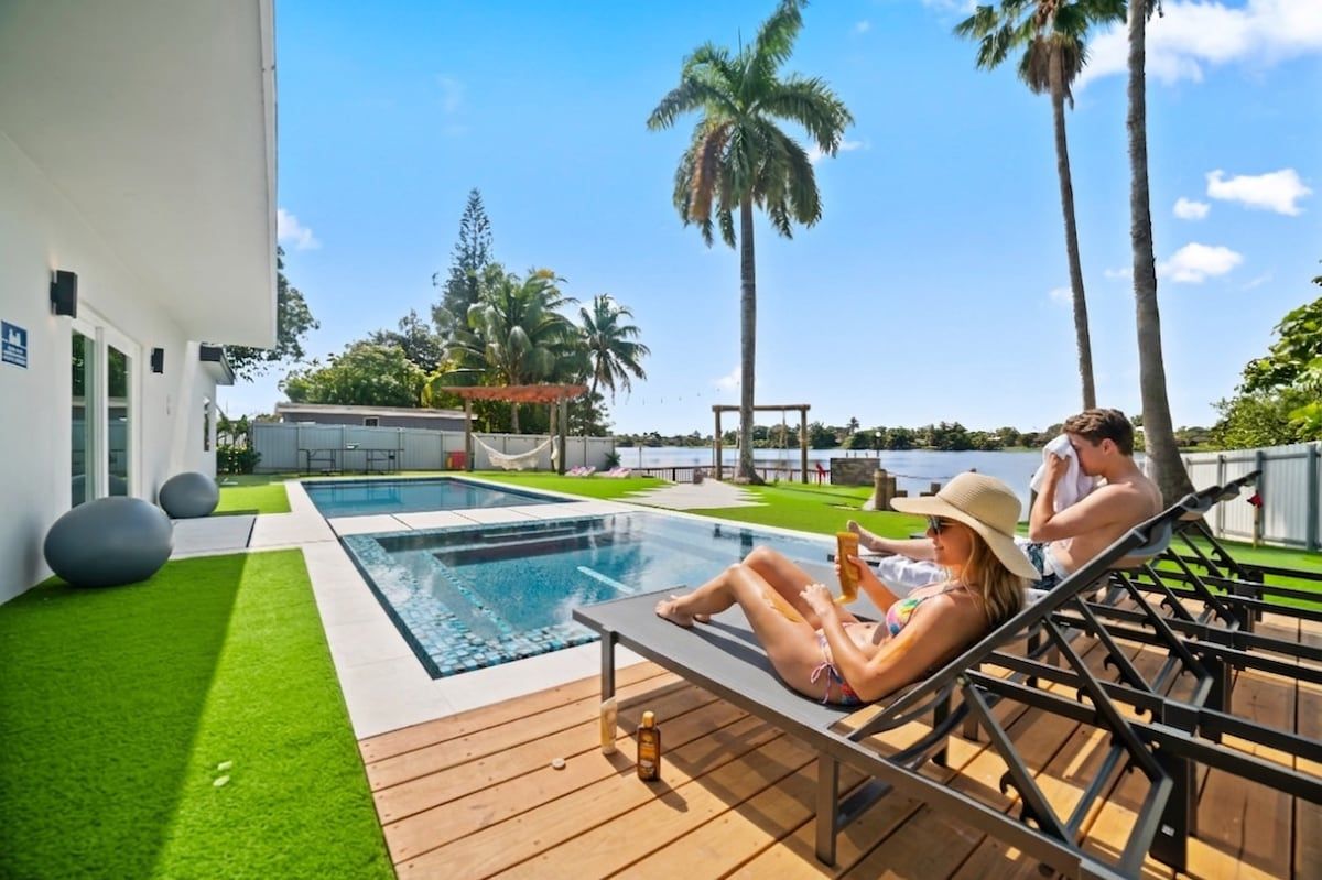 Ett område vid poolen med utsikt över vattnet, en terrass med solstolar och en byggnad med vit fasad.