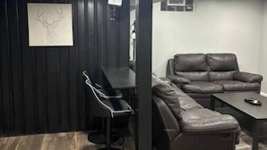 A modern living room with a dark wood floor, a black leather sofa, a black leather couch, a black dining table with four chairs, and a wall-mounted artwork of a deer head.