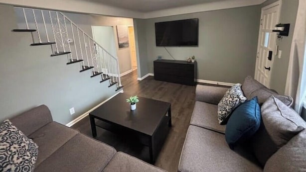 A living room with a staircase, a flat-screen TV, a dark entertainment unit, and a sectional sofa with patterned cushions.