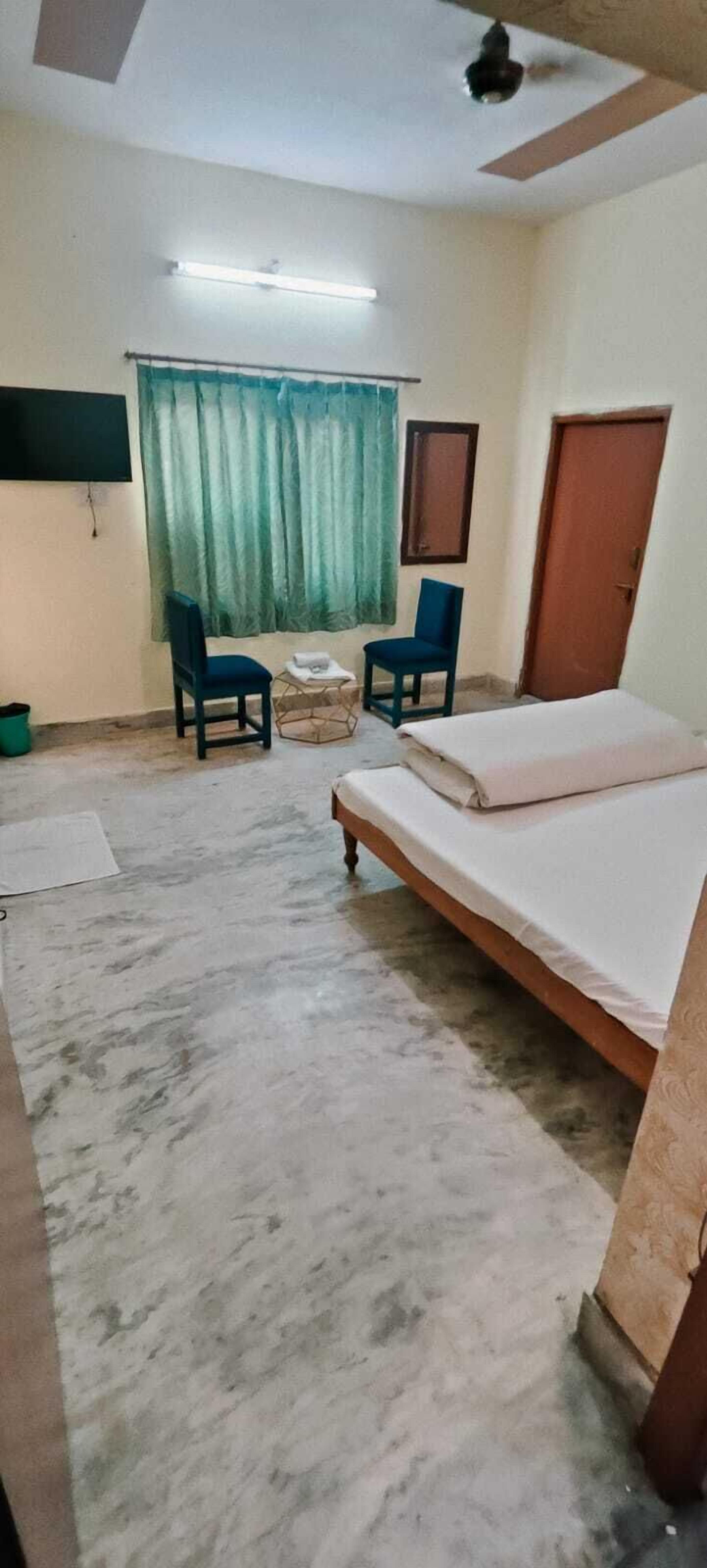 A room with a bed, two chairs, a small table, a window with curtains, a door, and a ceiling fan.