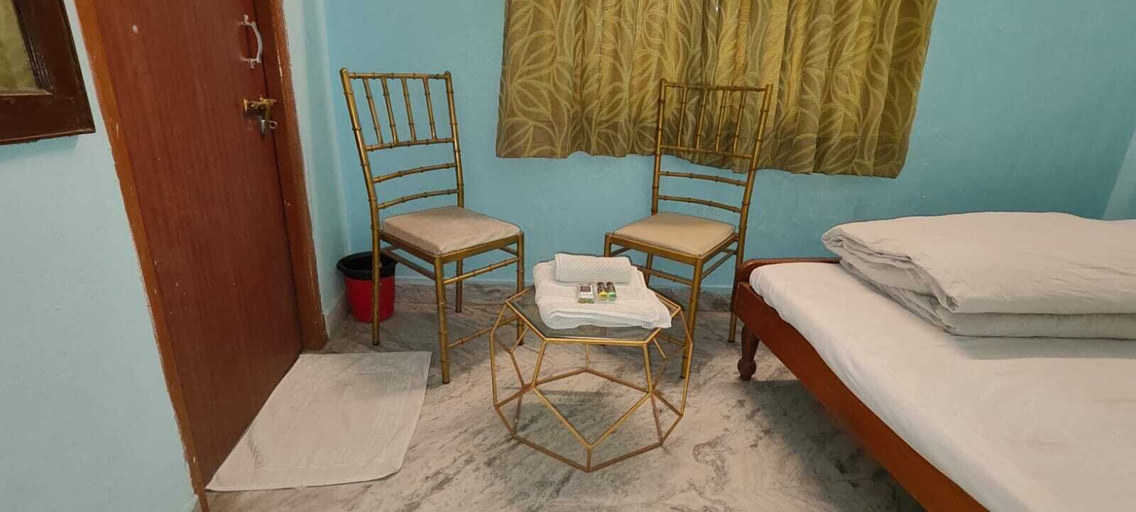 Una habitación pequeña y sencilla con cama, dos sillas, una mesita y ventana con cortina estampada.