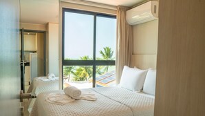 Hotelkamer met een groot raam, een bed met witte lakens, een badkamer met spiegel en handdoek, en uitzicht op palmbomen en zonnepanelen.