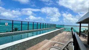 A rooftop infinity pool with a glass barrier, overlooking a beach and ocean.