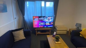 Een woonkamer met een flatscreen televisie waarop een Sky-kanaal wordt getoond, twee banken, een salontafel en een kleine tafel met een plant.