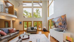 A spacious living room with a high ceiling, large windows, a fireplace, and a large TV.