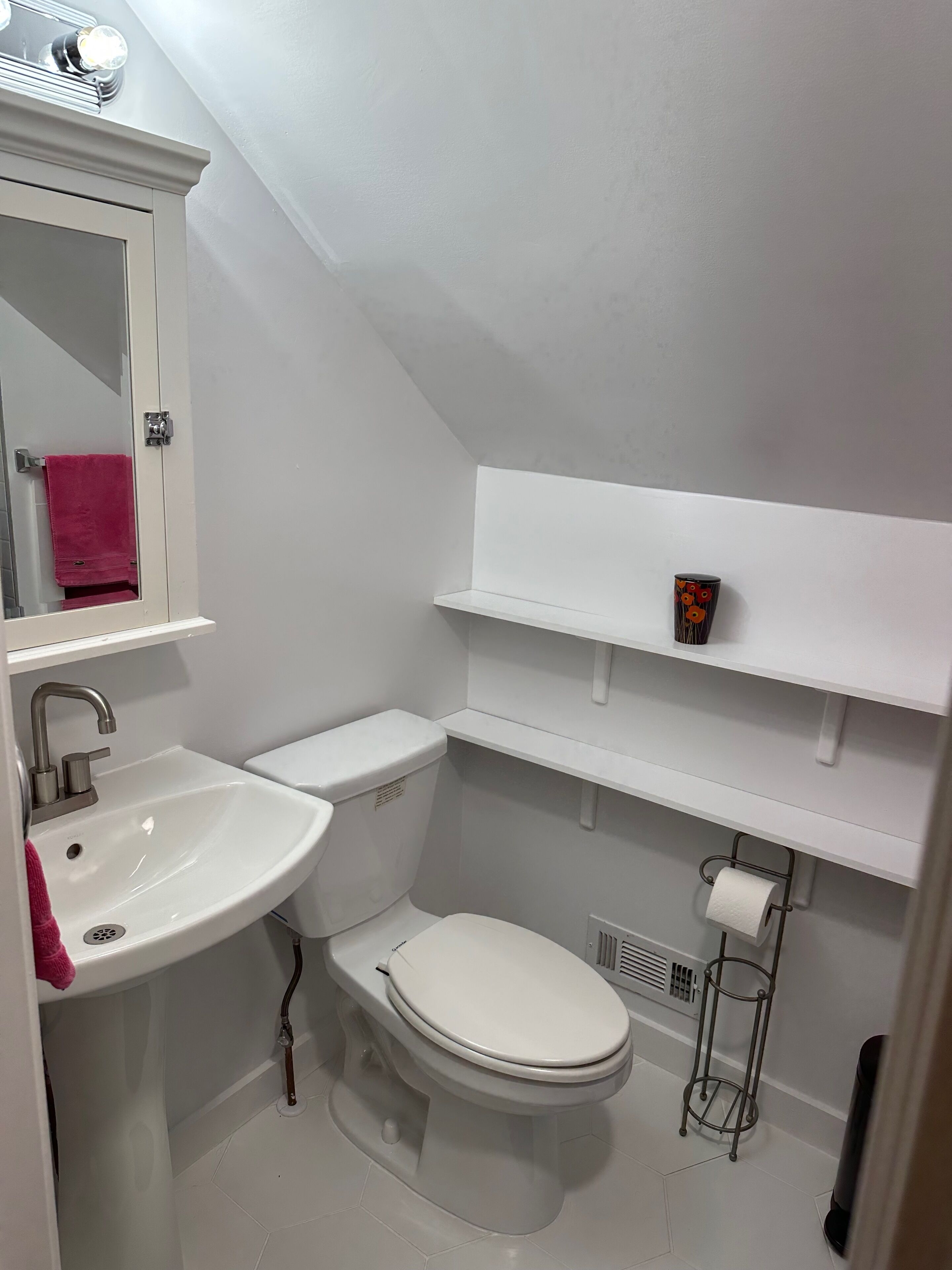 Un baño pequeño y blanco con lavamanos, inodoro y espejo. Hay estantes sobre el inodoro y una toalla colgada en el lavamanos.