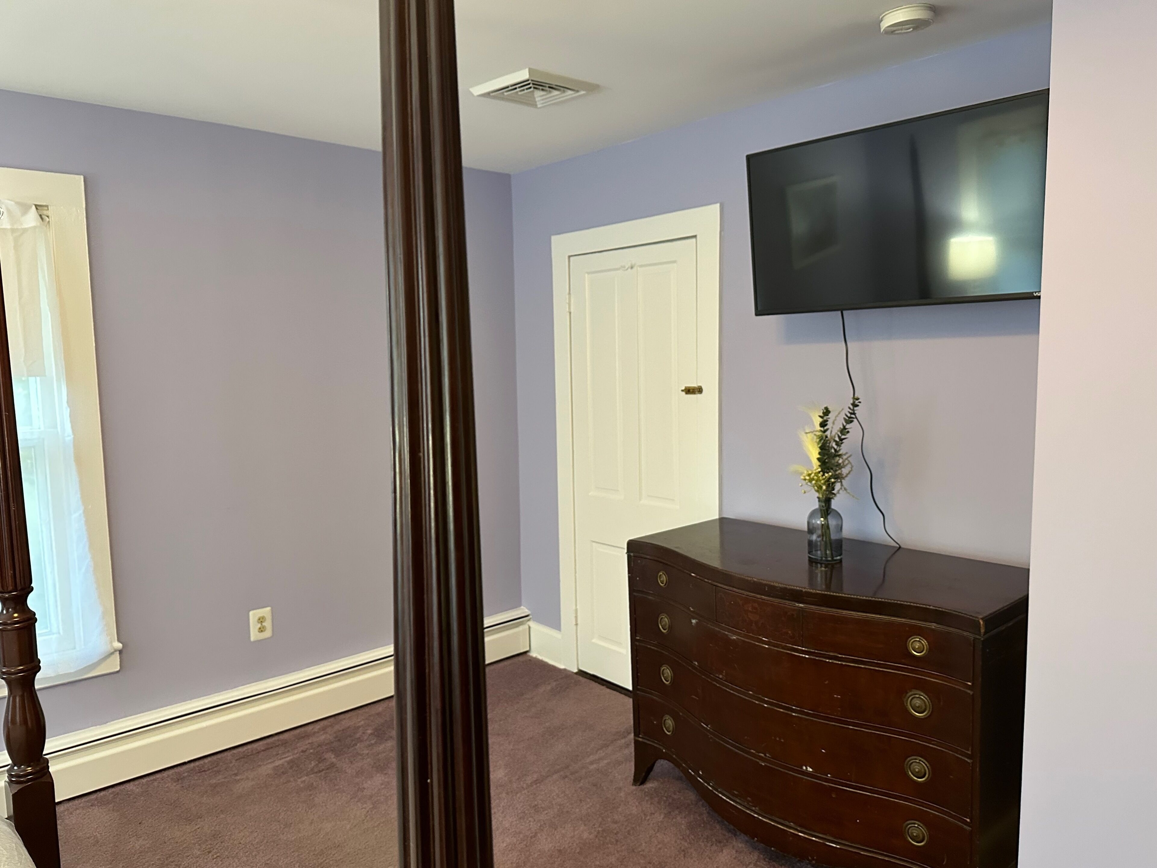 Une pièce avec une porte blanche, une commode en bois foncé, un téléviseur à écran plat accroché au mur et une fenêtre avec des stores.