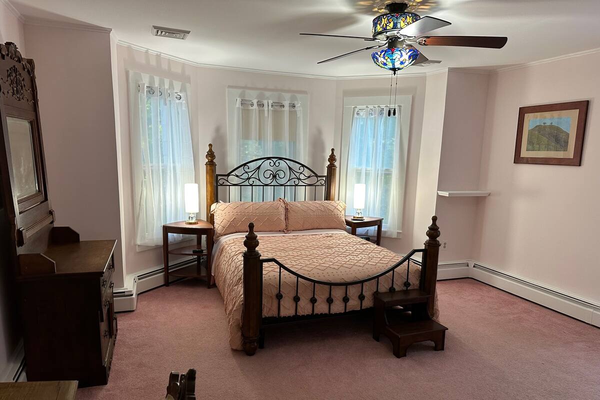 A bedroom with a four-poster bed, two bedside tables, a ceiling fan, and a window with sheer curtains.