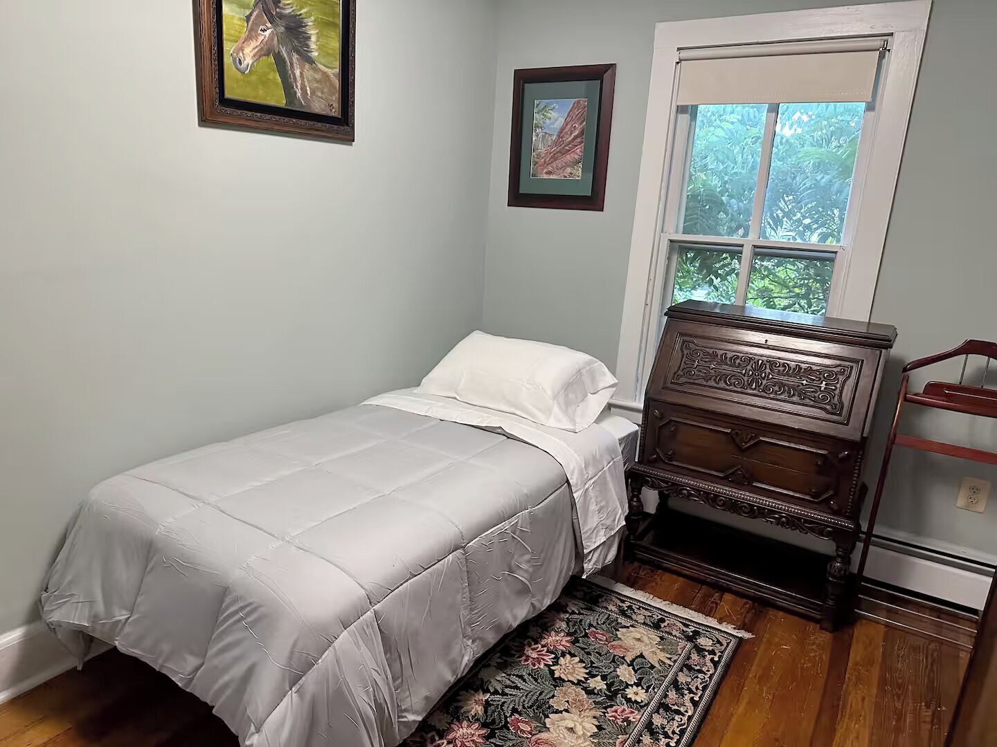 A bedroom with a single bed, a dresser, a window with a view of greenery, and two framed pictures on the wall.