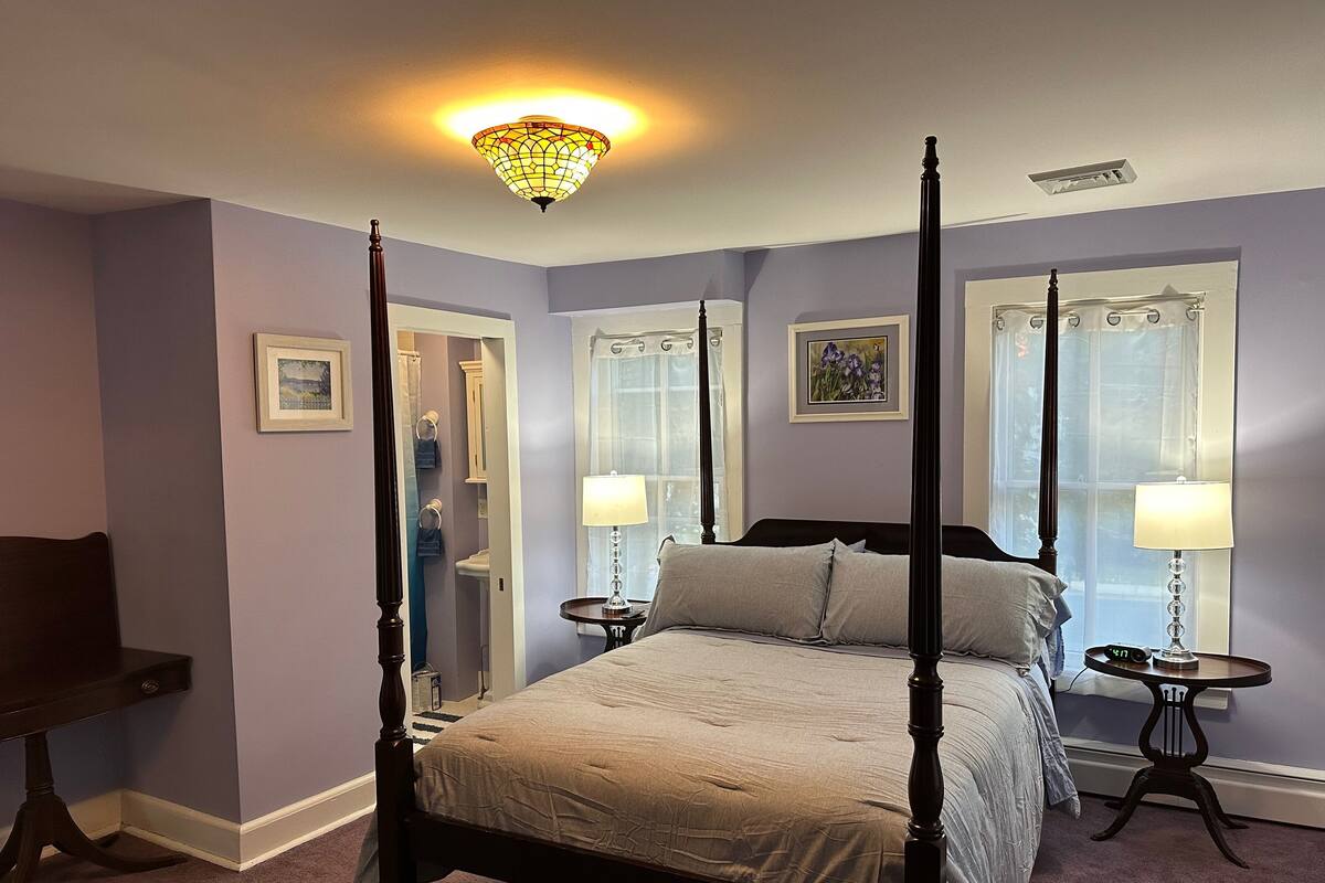 A four-poster bed with a beige bedspread, two bedside tables with lamps, a nightstand, a chair, and a ceiling light fixture.