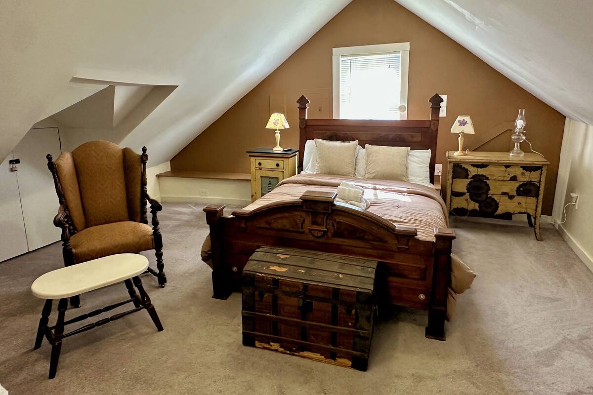 A bedroom with a sloped ceiling, a wooden bed with a headboard, two bedside tables with lamps, a wooden chair, and a chest of drawers.