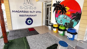 An outdoor area with a sign for 'Magarsus Elit Otel Restaurant', a painted mural of a van and palm trees, and waste bins labeled 'Kagit Atik Paper Waste'. - Magarsus Elit Hotel (Karatas)