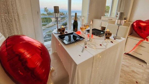 A dining table set with candles, a bottle, and a heart-shaped balloon in a room with a large window and a mirror. - Magarsus Elit Hotel (Karatas)