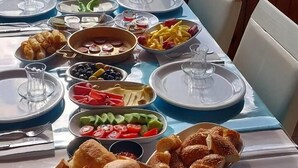 A table set for breakfast with various dishes, including croissants, fruits, and bread, with a view of the sea and buildings in the background. - Magarsus Elit Hotel (Karatas)