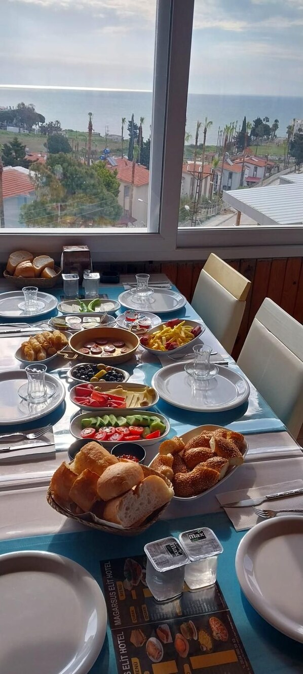 Una mesa dispuesta para el desayuno con diversos platos, incluyendo croissants, frutas y pan, con vista al mar y edificios en el fondo.
