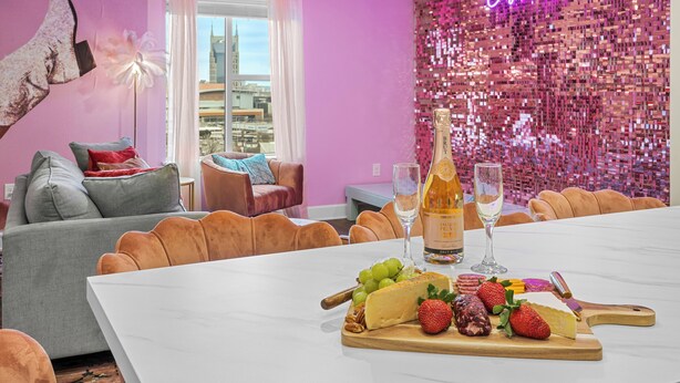 A modern living room with a large white table set for a meal, featuring a variety of cheeses, fruits, and a bottle of champagne.
