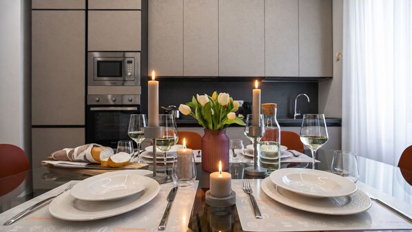Una mesa de comedor dispuesta con platos, cubiertos, vasos y velas, con un florero de flores y una botella de agua.