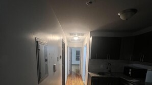 A narrow hallway with wooden flooring leading to a kitchen area with dark cabinets and a tiled backsplash.