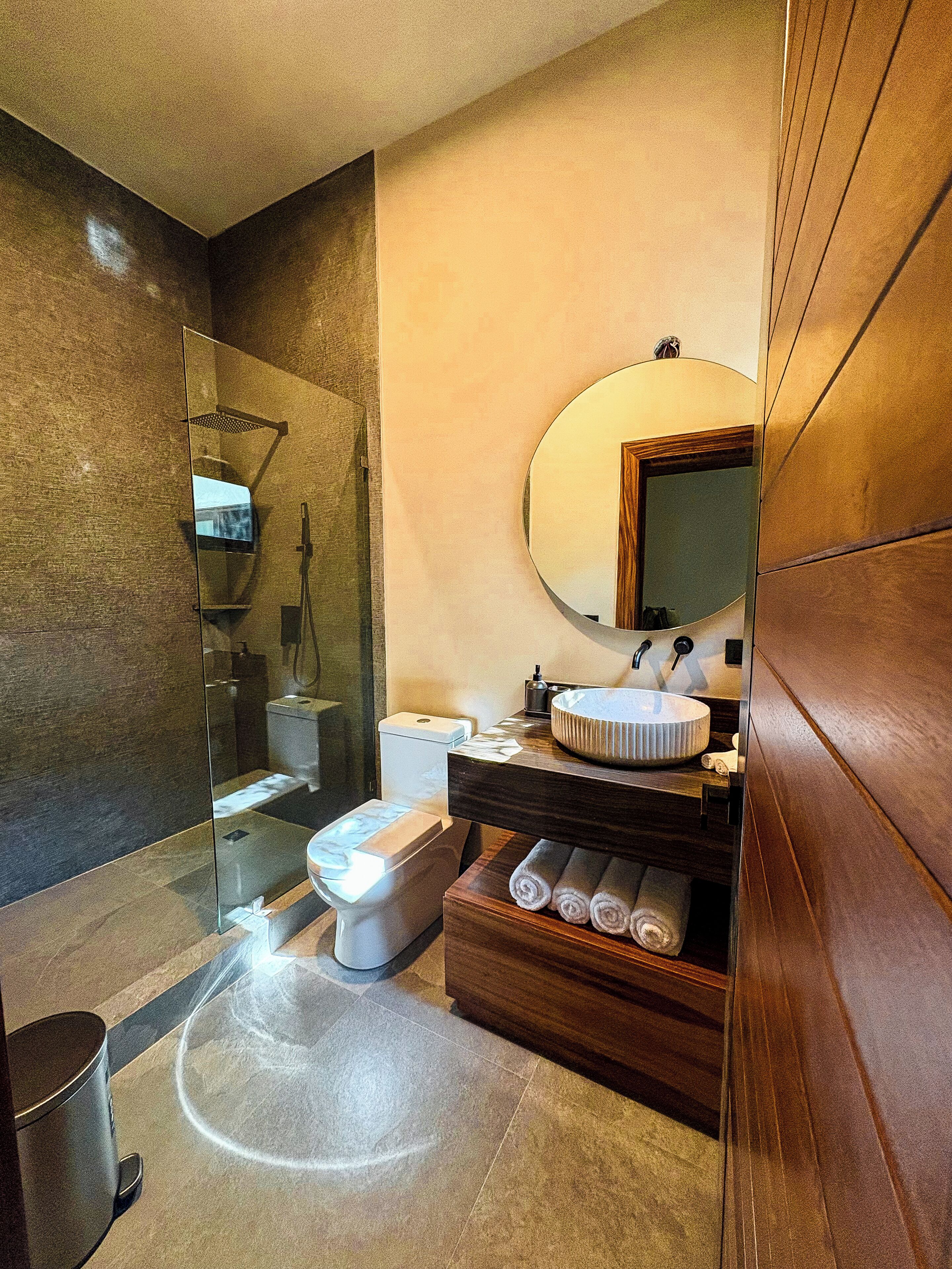 A modern bathroom with a glass-enclosed shower, a toilet, a sink with a round mirror, and neatly rolled towels.