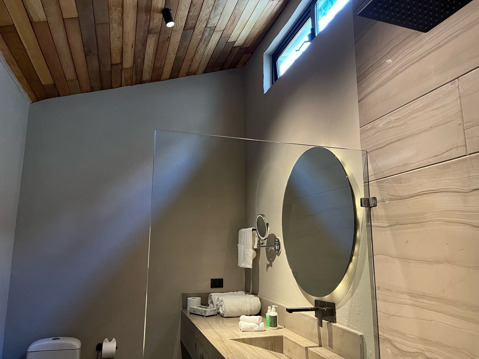 Un baño moderno con techo de madera, un espejo grande, un lavamanos con toallero y un inodoro.