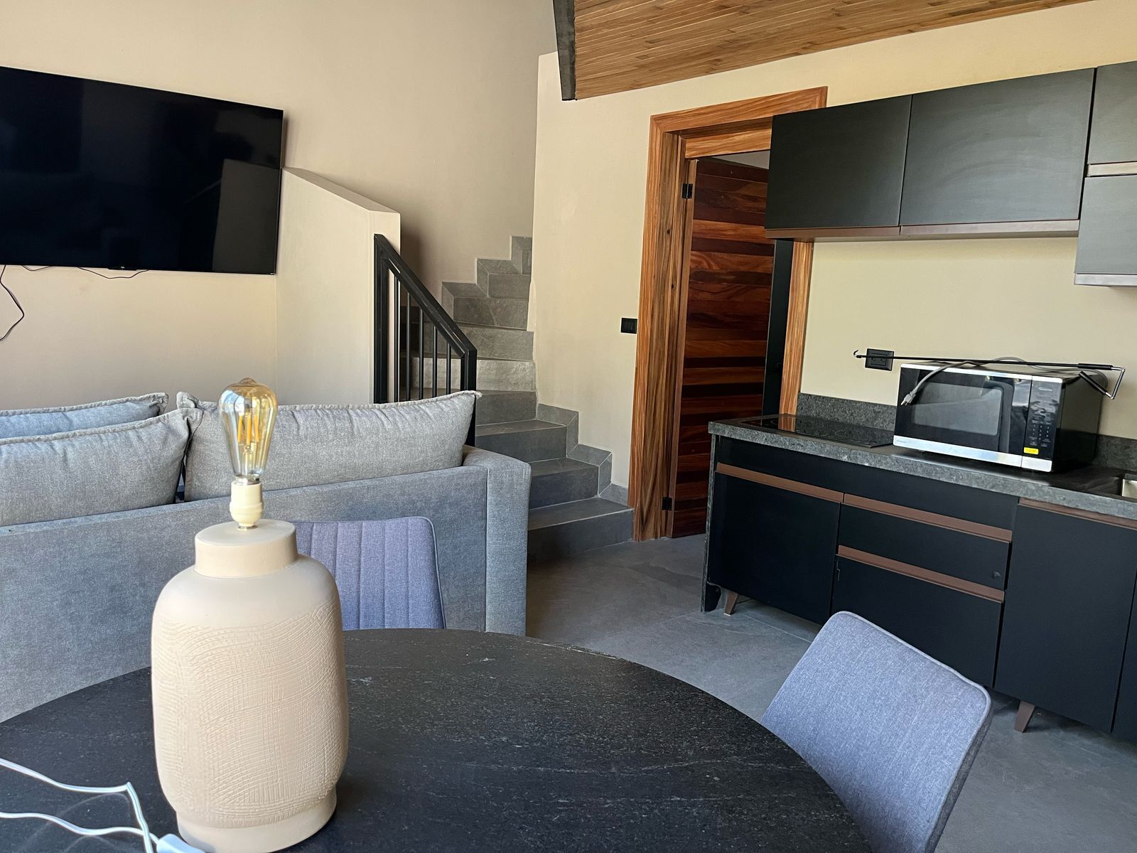 Een moderne woonkamer met een grijze bank, een ronde tafel met een lamp, een keuken met een magnetron, en een trap.