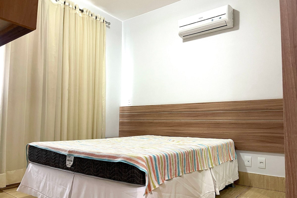 Une chambre à coucher avec un lit, une fenêtre avec des rideaux, un appareil de climatisation et une tête de lit en bois.