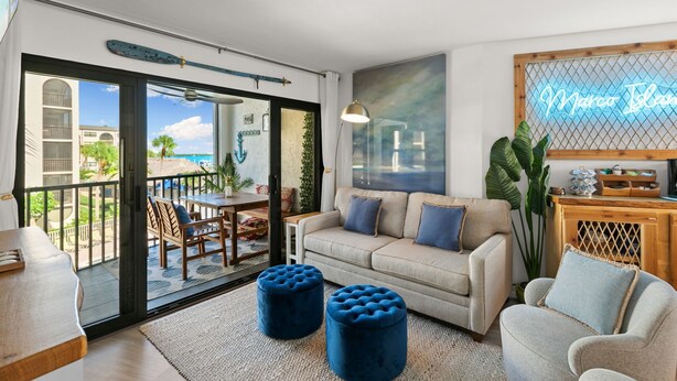 A living room with a beige sofa, two blue ottomans, a wooden side table, and a neon sign reading "Marco Island".