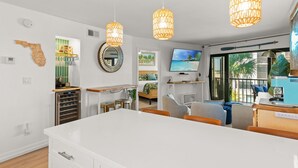 A modern kitchen with white cabinetry, a central island, and pendant lighting. There is a dining area with a large table and chairs, and a living area with a TV and comfortable seating.