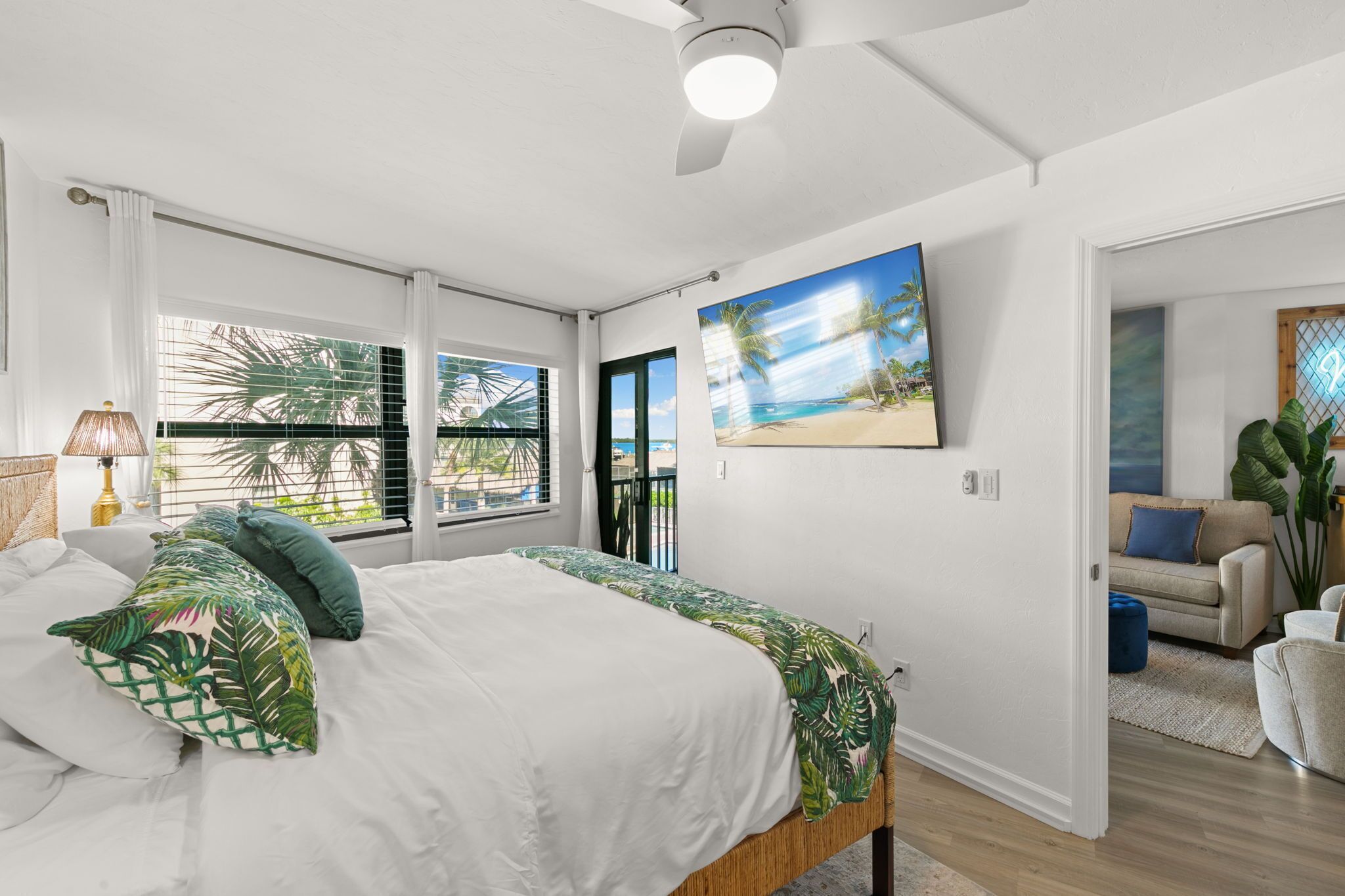 Un dormitorio con cama, ventana con persianas, un cuadro de playa y vista a palmeras.