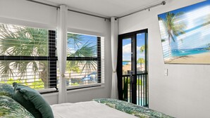 A bedroom with a bed, a window with blinds, a balcony door, and a beach view painting.