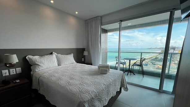 Ein modernes Hotelzimmer mit einem großen Bett, einem Nachttisch, einer Lampe und Blick auf das Meer und die Stadt vom Balkon aus.