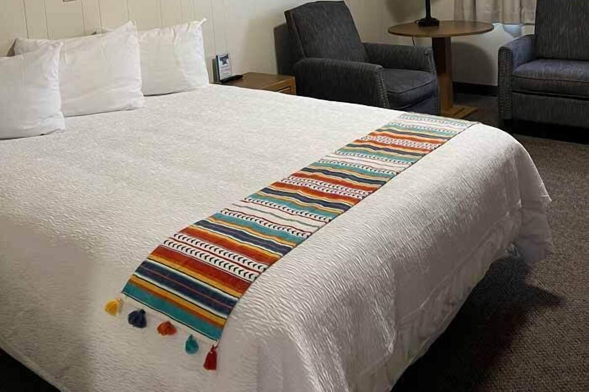 A hotel room with a large bed, two armchairs, a small table, and a wall sign saying "Be Our Guest".