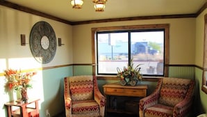 A room with two patterned armchairs, a wooden side table with a vase of flowers, a large window with a view of the outside, and a wall-mounted decorative plate. - Western Inn (Tremonton)