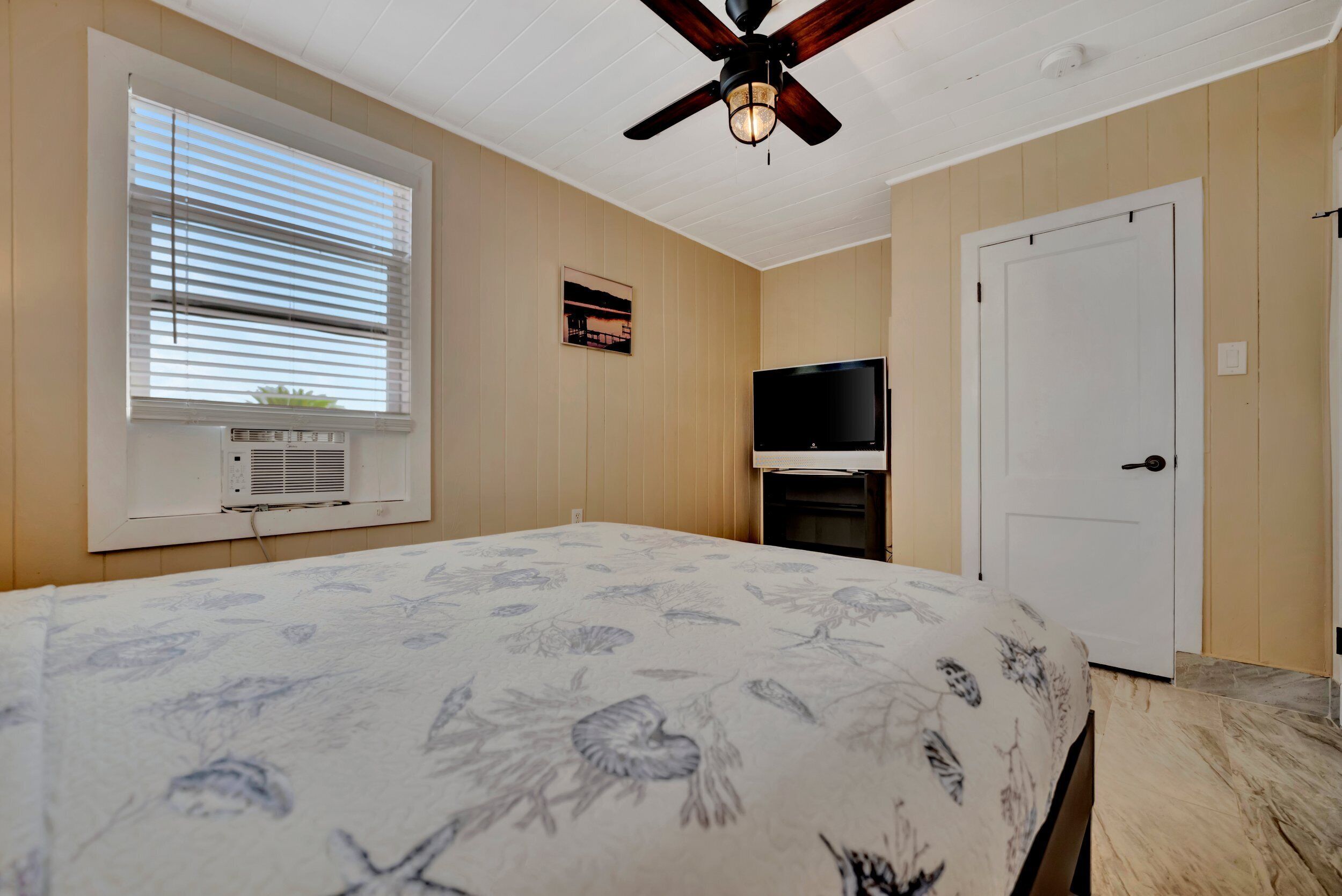 A bedroom with a bed, a ceiling fan, a TV, a door, and a window with blinds.