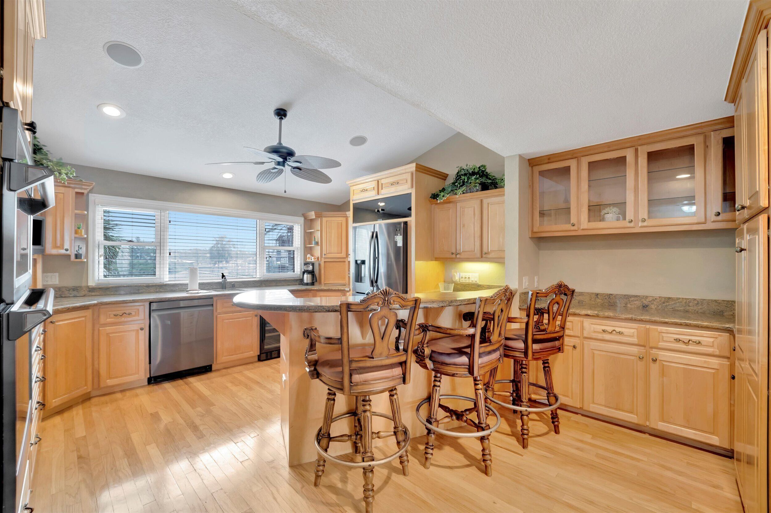 A modern kitchen with wooden cabinets, a central island with bar stools, stainless steel appliances, and a ceiling fan.