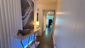 Un pasillo con suelo de madera oscura, una pared blanca con una obra de arte enmarcada, una mesa con lámpara y una puerta al final.