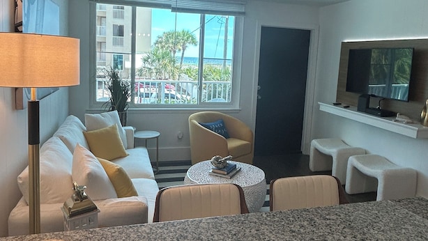 A modern hotel room with a white sofa, a small round table, a TV mounted on the wall, and a balcony with a view of the city.
