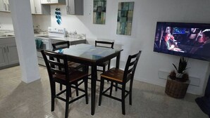 A dining area with a glass-top table and four chairs, a kitchen in the background, and a television on the wall. - Charming 2- Bedroom Apartment with AC, WiFi in St Thomas' East End (St Thomas)