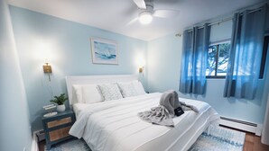 A bedroom with a bed, a nightstand, a lamp, a framed picture, and a window with curtains.