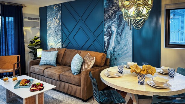 A modern living room with a brown sofa, a dining table set with plates and glasses, and a blue accent wall with a geometric pattern.