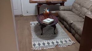 Uma sala de estar com sofá de couro marrom, mesa de centro redonda de madeira com um livro e uma cesta, tapete estampado e uma janela com persianas.