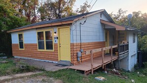 Uma casa pequena, térrea, com porta e janelas amarelas, varanda e antena parabólica.