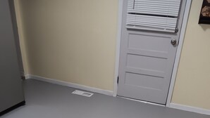Um quarto com piso claro, porta branca com janela e um pequeno respiradouro no chão.