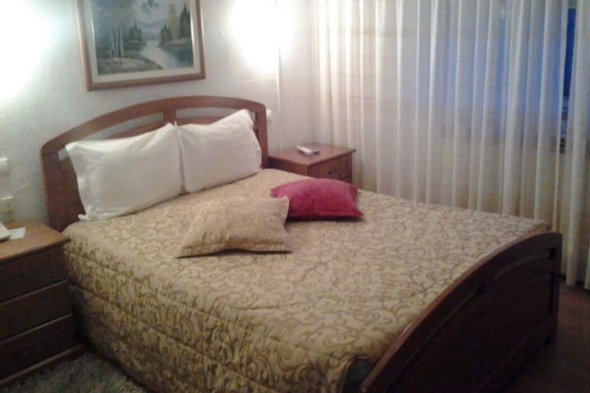 A neatly made bed with a patterned bedspread and pillows, a nightstand with a remote control, and a painting on the wall.