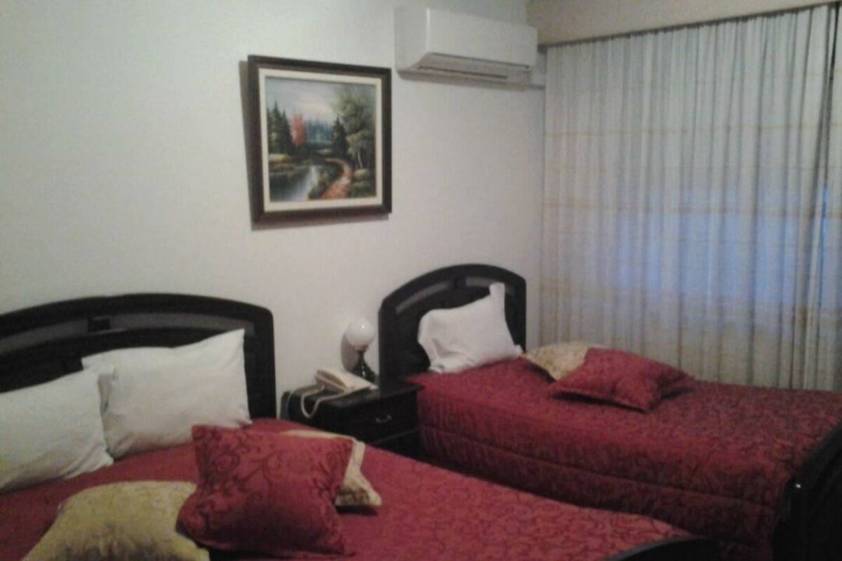 A hotel room with two beds, each with red bedding and multiple pillows. There is a painting on the wall and an air conditioning unit above the beds.