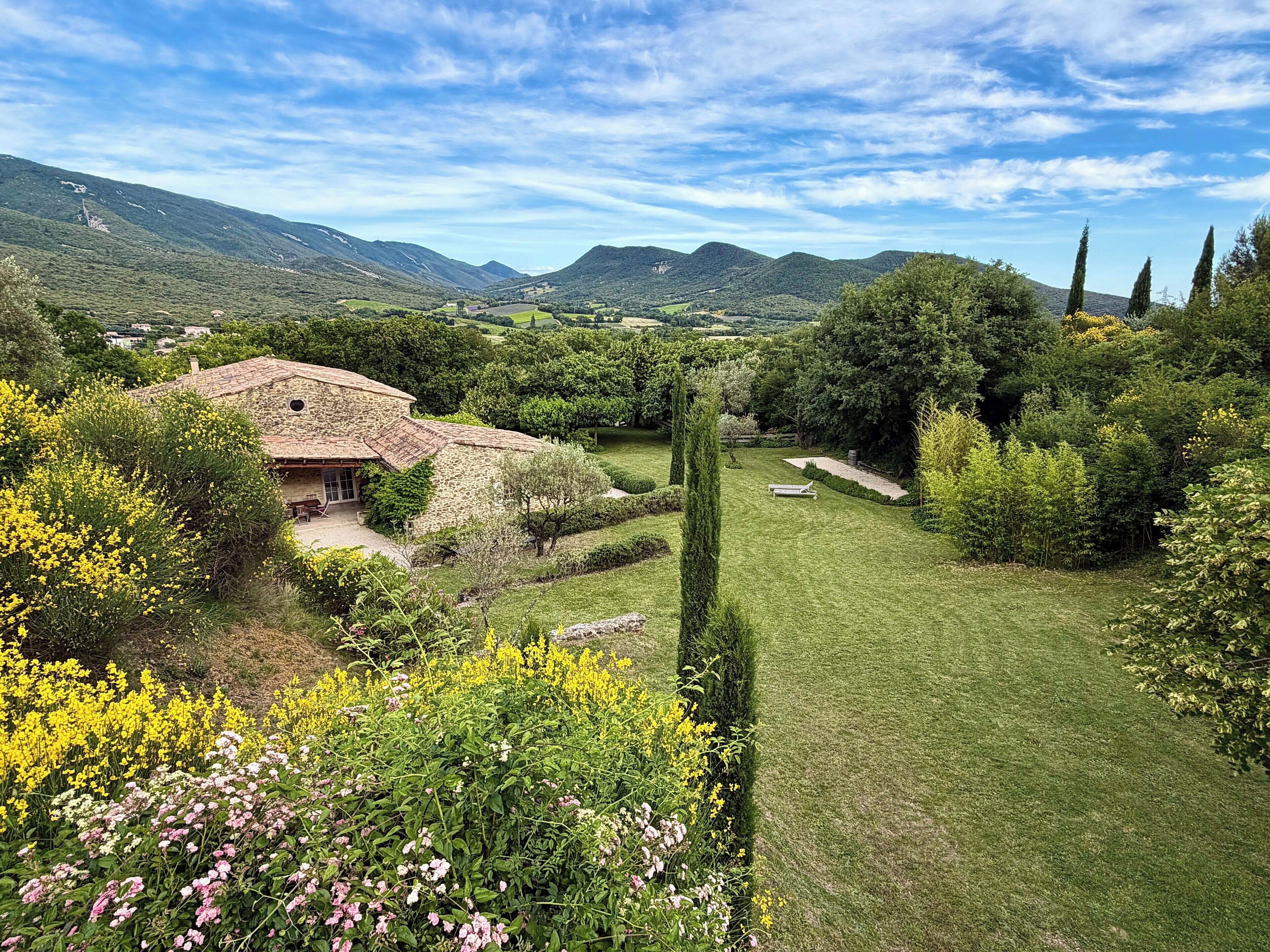 Une maison en pierre avec un toit en tuiles, entourée d’un jardin bien entretenu où poussent différentes espèces de plantes et où s’étend une pelouse.