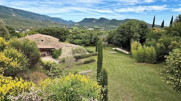 Une maison en pierre avec un toit en tuiles, entourée d’un jardin bien entretenu où poussent différentes espèces de plantes et où s’étend une pelouse.