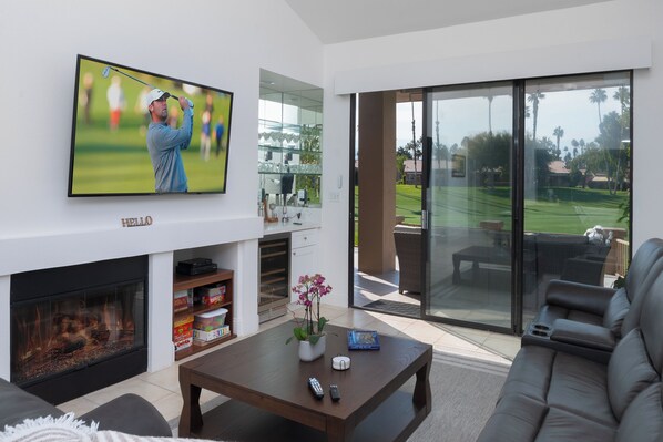 A modern living room with a flat-screen TV, a fireplace, a coffee table, and a large sliding glass door leading to a patio with outdoor furniture.
