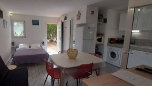 A compact room with a bed, a dining table with chairs, a washing machine, and a small kitchen area. - Mini-villa 2 pers. 3★ with garden, Residence with swimming pool - Near beaches (Lecci)